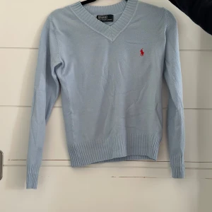 Ljusblå v-ringad tröja Polo Ralph Lauren - Supersnygg ljusblå v-ringad tröja från Polo Ralph Lauren med klassisk röd broderad logga på bröstet. Tröjan är stickad i mjukt material, har långa ärmar och breda ribbade muddar vid ärmslut och nederkant. .