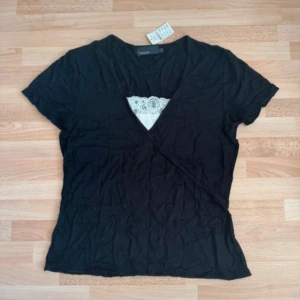 Ny. Svart omlottopp med spetsdetalj - Svart omlottopp från Kappahl med korta ärmar och snygg v-ringning. Toppen har en vit spetsdetalj vid urringningen som ger en extra touch. Perfekt att styla med jeans eller kjol för en trendig look.