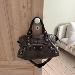 Balenciaga svart city skinnväska med detaljer - Den klassiska city väskan från Balenciaga i mjukt skinn med silverfärgade nitar, spännen och dragkedjor. Väskan har både handtag och avtagbar axelrem, samt flera fack och Mac 13 får plats i den. Köpt från collectors cage i 2023 för ca 7000 sek