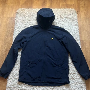 Mörkblå vindjacka från Lyle & Scott - Snygg svart vindjacka från Lyle & Scott med huva och dragkedja framtill. Jackan har det klassiska gula fågel-loggan på bröstet och två fickor med knappar. Perfekt för blåsiga dagar och enkel att matcha med allt.