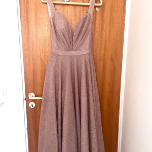 Glittrig champagnefärgad långklänning - Supersnygg långklänning i champagnefärg med glittrigt tyg och djup v-ringning. Klänningen har breda axelband, markerad midja och ett svepande, utsvängt kjolparti. Perfekt för bal eller fest där du vill sticka ut och glänsa.