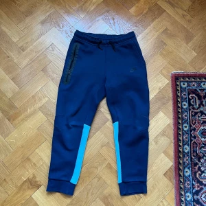 Nike tech blå mjukisbyxor med ljusblå detaljer - Snygga mörkblå mjukisbyxor från Nike med ljusblå paneler på benen. Byxorna har resår och snörning i midjan, dragkedjeficka på ena sidan och ribbade muddar vid bensluten. Perfekta för chill eller träning.