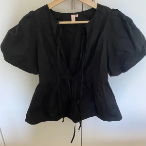 Svart blus med puffärm från Nelly - Supersnygg svart blus från Nelly med puffiga korta ärmar och knytband framtill. Blusen har en lätt v-ringning och markerad midja som ger en fin siluett. Perfekt för dig som gillar trendiga detaljer och en feminin look.