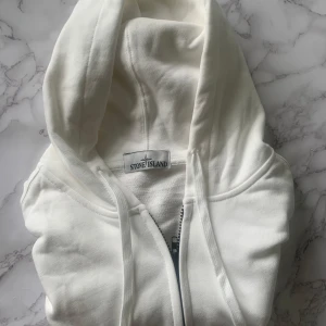 Vit hoodie från Stone Island - Säljer en clean vit hoodie från Stone Island med dragkedja och klassisk huva. Tröjan har långa ärmar, snörning i huvan och den ikoniska avtagbara loggan på ärmen. Tillverkad i mjuk bomull, perfekt för en chill och stilren look.