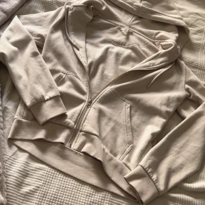 Beige/vit  hoodie med dragkedja XS - Säljer en beige hoodie i storlek XS med dragkedja framtill och huva. Tröjan har långa ärmar, ribbade muddar och två fickor framtill. Perfekt för en chill och avslappnad stil. Materialet känns mjukt och skönt, bomull och polyester 