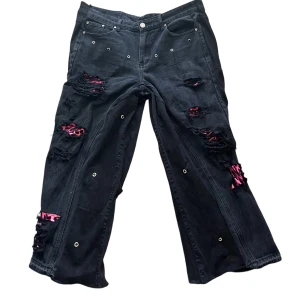 Custom svarta wide jeans med rosa detaljer - Unika svarta jeans med bred passform, slitna detaljer och rosa leopardmönstrat tyg under trasorna. Byxorna har metallöljetter och klassisk femficksdesign. Perfekta för dig som vill sticka ut med en edgy streetstil. Hör av dig om du vill ha måtten. 