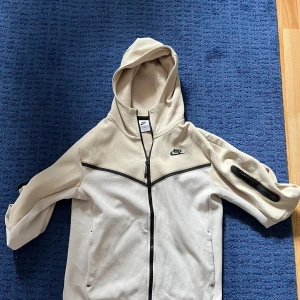 Nike beige och vit hoodie, tech fleece S - Nike hoodiejacka i beige och vitt med svart logga på bröstet. Jackan har hel dragkedja, huva och en praktisk ficka med dragkedja på ärmen. Tillverkad i mjuk bomullsmix, perfekt för chill eller träning. Snygg kontrast med svarta detaljer och sportig look. Helt ny utan användning. 