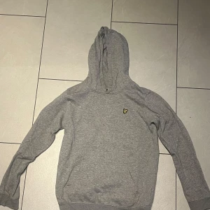 Grå hoodie från Lyle & Scott - Snygg grå hoodie från Lyle & Scott med klassisk gul logga på bröstet. Tröjan har huva, magficka och långa ärmar. Perfekt för en chill och avslappnad stil. Mjuk och skön bomullskänsla.