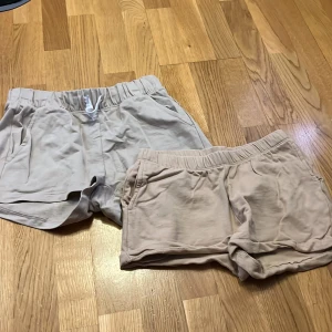 Beige bomullsshorts H&M NYC, stl 158 - Två par mjuka och sköna shorts i beige från H&M, båda i ekologisk bomull. Ena paret har broderad NYC-logga och vit snörning i midjan, båda har elastisk midja och fickor. Perfekta för chill dagar eller träning under sommaren.