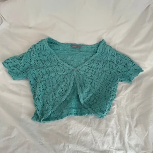 Turkos stickad kofta från KappAhl - Supersöt turkos stickad kofta från KappAhl med kort ärm och v-ringning. Koftan har ett genombrutet spetsmönster och knäpps framtill med två små knappar. Croppad modell som passar perfekt till sommarens outfits.