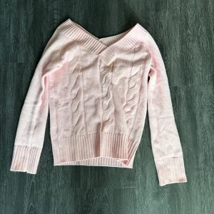 Ljusrosa stickad offshoulder tröja - Supersöt ljusrosa stickad tröja med offshoulder och bred ribbad kant vid halsen. Tröjan har kabelstickat mönster framtill och långa ärmar. Perfekt för dig som vill ha en feminin och trendig look med en mysig vibe.