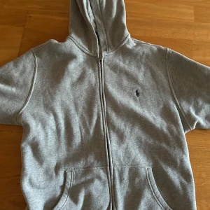Polo Ralph Lauren.   - kommer från kids brandstore. Klassisk grå hoodie från Polo Ralph Lauren med dragkedja och huva. Tröjan har broderad logga på bröstet och två fickor framtill. Perfekt för chill dagar och enkel att matcha med jeans eller joggers.