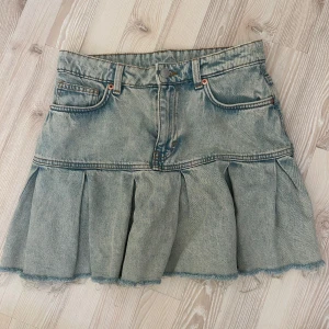 Jeanskjol med volang från Monki - Säljer en ljusblå jeanskjol från Monki med volang nertill och rå kant. Kjolen har klassiska jeansfickor, hällor för bälte och en snygg plisserad detalj som ger extra volym. Aldrig använd.