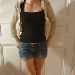 Beige stickad bolero - Säljer en beige stickad bolero från KappAhl med snyggt hålmönster och vågig kant. Västen har trekvartsärmar, en knapp framtill och är croppad i modellen. Perfekt för lager-på-lager och ger en chill, boho-inspirerad vibe. Köpt på en second hand men har personligen aldrig använt då den inte riktigt passar min stil längre, men ändå så snygg och i bra skick!❤️
