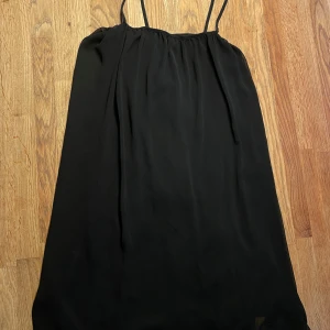 Svart kort klänning - Svart strechig kort klänning med tunna axelband från JC. Klänningen har en svart tajt innerklänning fäst under det svarta lite genomskinliga lagret med lös passform. Använd endast en gång! Den har också en snygg rosett i ryggen som man även kan knyta runt midjan om man hade velat det!🖤