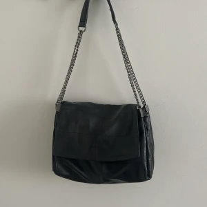 Svart axelväska i skinn med kedja från Zara - Snygg svart axelväska i mjukt skinn med bred lockstängning och coola silverfärgade kedjor som axelrem. Väskan har en stilren och enkel design som passar till det mesta. Perfekt storlek för att få plats med allt du behöver till vardags. Populär väska från Zara