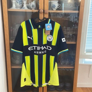Manchester City bortaställ 2024-2025  Puma M - Manchester Citys officiella matchtröja från Puma i storlek M. Tröjan har svart bas med neon-gula och blå vertikala ränder, klubbmärke på bröstet och Etihad Airways-tryck. Tillverkad i tekniskt DryCell-material som andas. Kortärmad och perfekt för fotbollsfans. Jag själv är en stor fotbollströjsamlare och jag garanterar att allt jag säljer är 100% äkta vilket syns i bilderna!