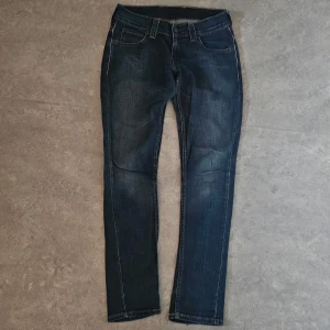 Levis 501 mörkblå jeans straight fit - Klassiska Levis 501 jeans i mörkblå tvätt med raka ben och femficksmodell. Snygga slitningar och tydliga sömmar ger en cool vintagekänsla. Ikonisk Levis-lapp bak i midjan och röd etikett på bakfickan.