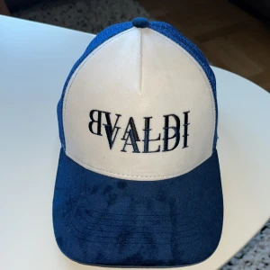 Blå och vit keps från BVALDI - Snygg truckerkeps från BVALDI med vit frontpanel och blått broderat logotyp. Skärmen och baksidan är blå, med meshmaterial för extra ventilation. Justerbar passform och cool kontrast mellan färgerna. Perfekt accessoar för en streetwear-look. Använd en gång och ser helt ut som nyskick.