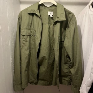 Olivgrön overshirt från H&M - Olivgrön overshirt från H&M med klassisk krage, dragkedja framtill och två bröstfickor med lock. Jackan är tillverkad i bomull och har en avslappnad passform som passar perfekt till lager-på-lager-stil.