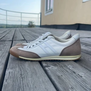Hogan Rebel sneakers i vitt och beige - Snygga Hogan Rebel sneakers i vitt skinn med beige mockadetaljer och bruna gummisulor. Klassisk retrostil med rund tå, snörning och diskret H-logga på sidan. Perfekta för dig som gillar stilrena och sportiga skor med premiumkänsla.