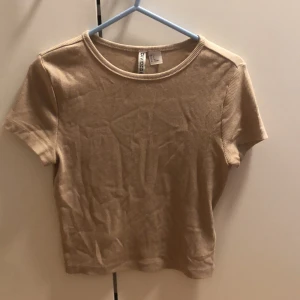 Beige ribbad topp från H&M Divided - Kortärmad ribbad topp i beige från H&M Divided. Modellen är croppad med rund halsringning och har en enkel, stilren look som passar perfekt till jeans eller kjol. Mjuk och stretchig bomullskvalitet för en bekväm känsla.