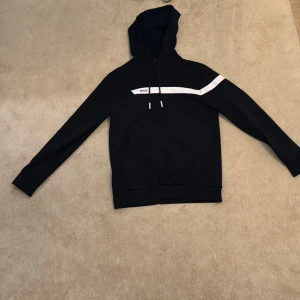 Svart hoodie från BOSS med vit detalj - Svart hoodie från BOSS med vit rand över bröstet och på ena ärmen. Klassisk huva med snörning och långa ärmar. Snygg och stilren design som passar perfekt till jeans eller joggers. Har haft på mig den max 4-5 gånger. Har haft den i 2 månader. Pris kan diskuteras vid sänkning. Storlek S men M kan också ta på sig den, den är lite stor i storlek. 