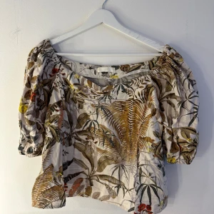 Blommig blus från H&M  - Supersöt blommig blus från H&M i storlek L. Toppen har puffiga trekvartsärmar, bred resår vid halsen och ett luftigt material av linne och viskos. Mönstret går i beige, brunt, grönt och inslag av gult och rött. Perfekt för en chill och trendig look.