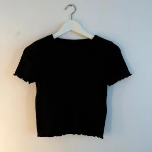 Svart ribbad topp från Gina Tricot - En svart, croppad ribbad topp från Gina Tricot med korta ärmar och vågiga kanter vid ärmslut och nederkant. Enkel och stilren modell som är lätt att matcha med allt. Perfekt för dig som gillar en clean look med lite edge. Storlek s