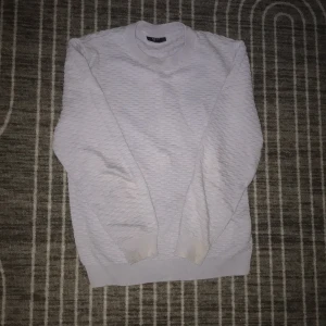 Vit strukturerad tröja från SMOG - Vit långärmad tröja från SMOG i regular fit. Tröjan har ett diskret strukturerat mönster och ribbade muddar vid ärmslut och nederkant. Perfekt för dig som gillar en clean och enkel stil med lite extra textur.