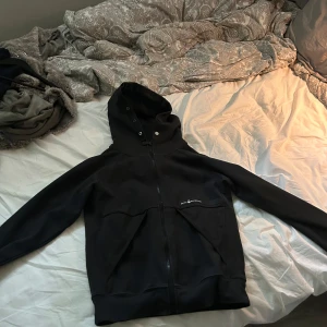 Svart vindjacka från Endurance - Svart vindjacka från Endurance med huva och hel dragkedja framtill. Jackan har diskret logga på bröstet, två stora fickor och meshfoder på insidan. Perfekt för blåsiga dagar och enkel att matcha med det mesta.