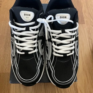 Svarta Dior B30 sneakers med vita detaljer - Dior B30 sneakers i svart mesh och syntet med vita och silverfärgade detaljer. Snygg CD-logga på sidan och bak, samt vita snören och en chunky vit sula. Sportig och modern look som sticker ut med kontrasterande paneler och cool design.