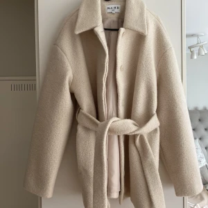 Beige kappa med bälte från NA-KD - Säljer en beige kappa från NA-KD i storlek 40. Jackan har en oversized passform, bred krage och knyts med bälte i midjan. Materialet är ullmix med en mjuk, lite fluffig yta. Perfekt för höst och vinter när du vill vara både varm och snygg.
