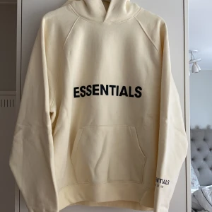 Essentials hoodie i beige från Fear of God - Snygg beige hoodie från Fear of God Essentials med svart logga på bröstet och ärmen. Oversized passform, stor huva och känguruficka framtill. Mjuk bomullskvalitet. 