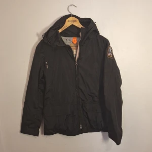 Svart vindjacka från Parajumpers M - Svart vindjacka från Parajumpers med huva och dragkedja framtill. Jackan har en klassisk patch på ärmen och orange detalj i nacken. Lättviktig och perfekt för mellansäsong. Snygg och funktionell design med flera fickor och justerbar huva.