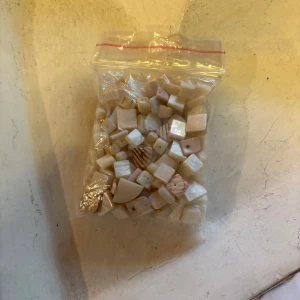 Pärlemorvita pärlor i olika former - En påse med pärlemorvita pärlor i olika former och storlekar, perfekta för att skapa egna smycken som armband, halsband eller örhängen. Pärlorna har en skimrande yta och vissa har mönster eller ränder. Passar dig som gillar DIY och unika accessoarer.