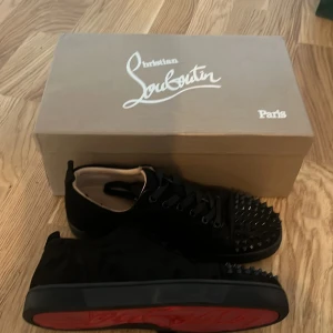 Svarta Christian Louboutins med nitar  - Säljer ett par svarta sneakers från Christian Louboutin med klassisk röd sula och svarta nitar på tån. Skorna har snörning och är tillverkade i mocka med läderdetaljer på insidan. Perfekta för dig som vill sticka ut med en edgy stil. Storleken är 39,5 och dom är större i storleken som passar 40,5