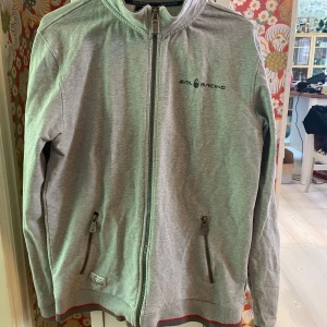 Grå zipjacka från Sail Racing L - Grå jacka från Sail Racing i storlek L. Jackan har lång ärm och logga på bröstet samt en patch på ärmen. Perfekt för dig som gillar sportig och clean stil.