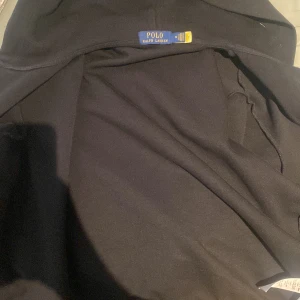 Svart hoodie tjocktröja  från Polo Ralph Lauren - Svart hoodiejacka från Polo Ralph Lauren med klassisk broderad logga på bröstet. Jackan har huva och är tillverkad i mjukt bomullsmaterial, perfekt för en chill och stilren look. Passar dig som gillar enkel och tidlös design.
