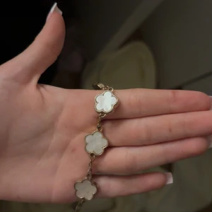 Guldigt armband med vita blommor - Superfint armband med flera blommor i vit pärlemor och guldfärgad metall. Blommorna har en mjuk, rundad form och sitter på rad längs kedjan. Perfekt accessoar för att lyfta din outfit med en söt och elegant touch.
