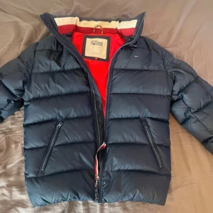 Mörkblå pufferjacka Tommy Hilfiger - Snygg mörkblå pufferjacka från Tommy Hilfiger Denim med röd insida och klassiska detaljer. Jackan har dragkedja framtill, två snedställda fickor med dragkedja och diskret logga på bröstet. Jättebra skick, endast använd ca 4-5 gånger