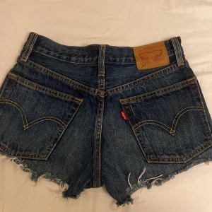 Levi's 501 jeansshorts med fransar - Snygga mörkblå jeansshorts från Levi's 501 med råa fransade kanter och slitningar. Klassisk femficksmodell, knäppning med knappar och tydliga kontrastsömmar. Perfekta för sommaren och ger en cool vintagekänsla.