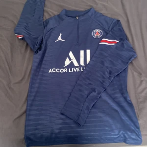 PSG långärmad träningströja Jordan - Mörkblå långärmad PSG träningströja från Jordan med halv dragkedja, vita loggor och diskret mönster. Röd, vit och blå rand på armarna.
