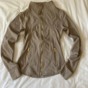 Lululemon defyne jacket - Supersnygg beige träningsjacka från Lululemon med guldfärgade dragkedjor och figurnära passform. Jackan har hög krage, långa ärmar och smarta fickor med dragkedja. Perfekt för gymmet eller powerwalks när du vill känna dig både sportig och stilren. Storlek 2 vilket motsvarar S/XS. Helt ny!