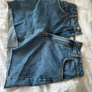 Blå jeansshorts från Bershka - Snygga blå jeansshorts från Bershka med råa, avklippta kanter och klassiska fem fickor. Shortsen har normal passform och dragkedja framtill. Perfekta för sommardagar och ger en avslappnad vibe.