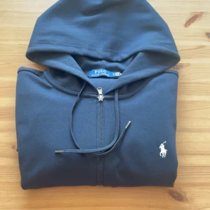 Svart hoodie från Polo Ralph Lauren - Säljer en svart hoodie från Polo Ralph Lauren med dragkedja och huva. Klassisk design med logga broderad på bröstet och fickor framtill. Perfekt för en avslappnad stil. Står L men passar för m