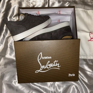 Grå mocka sneakers från Louboutin - Snygga grå sneakers i mocka från Christian Louboutin med klassisk vit sula och ikonisk röd undersida. Skorna har rund tå, snörning och stilren design som passar perfekt till streetwear eller en mer dressad look.