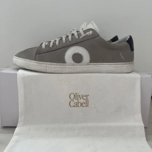 Oliver Cabell Low 1 sneakers grå 44 - Stilrena sneakers från Oliver Cabell i modellen Low 1. Skorna är gråa med vita detaljer, rund tå och vit sula. De har snörning och är tillverkade i skinn. Snygg cirkeldetalj på sidan och svart hälparti. Perfekta för dig som gillar minimalistisk design. Skorna är andvända sparsamt. Dustbag & box medföljer!Skriv bara om ni har några frågor eller funderingar!