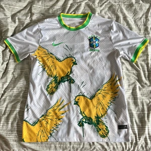 Vit Brasilien fotbollströja Nike - Snygg vit Brasilien fotbollströja från Nike med stora gula fåglar och gröna detaljer. Tröjan har korta ärmar, grönt och gult runt hals och ärmslut samt Brasilien-emblem på bröstet. Perfekt för dig som vill sticka ut på match eller i vardagen.