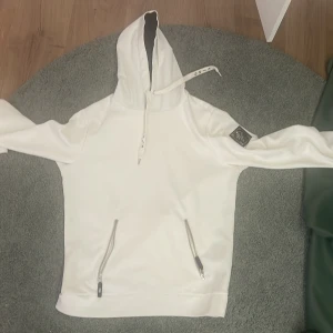 Vit hoodie med dragkedjor och patch märke goja - Snygg vit hoodie med huva och snörning, två dragkedjefickor framtill och en svart patch på ena ärmen. Hoodien har lång ärm och en clean look, perfekt för dig som gillar stilrena plagg med coola detaljer.